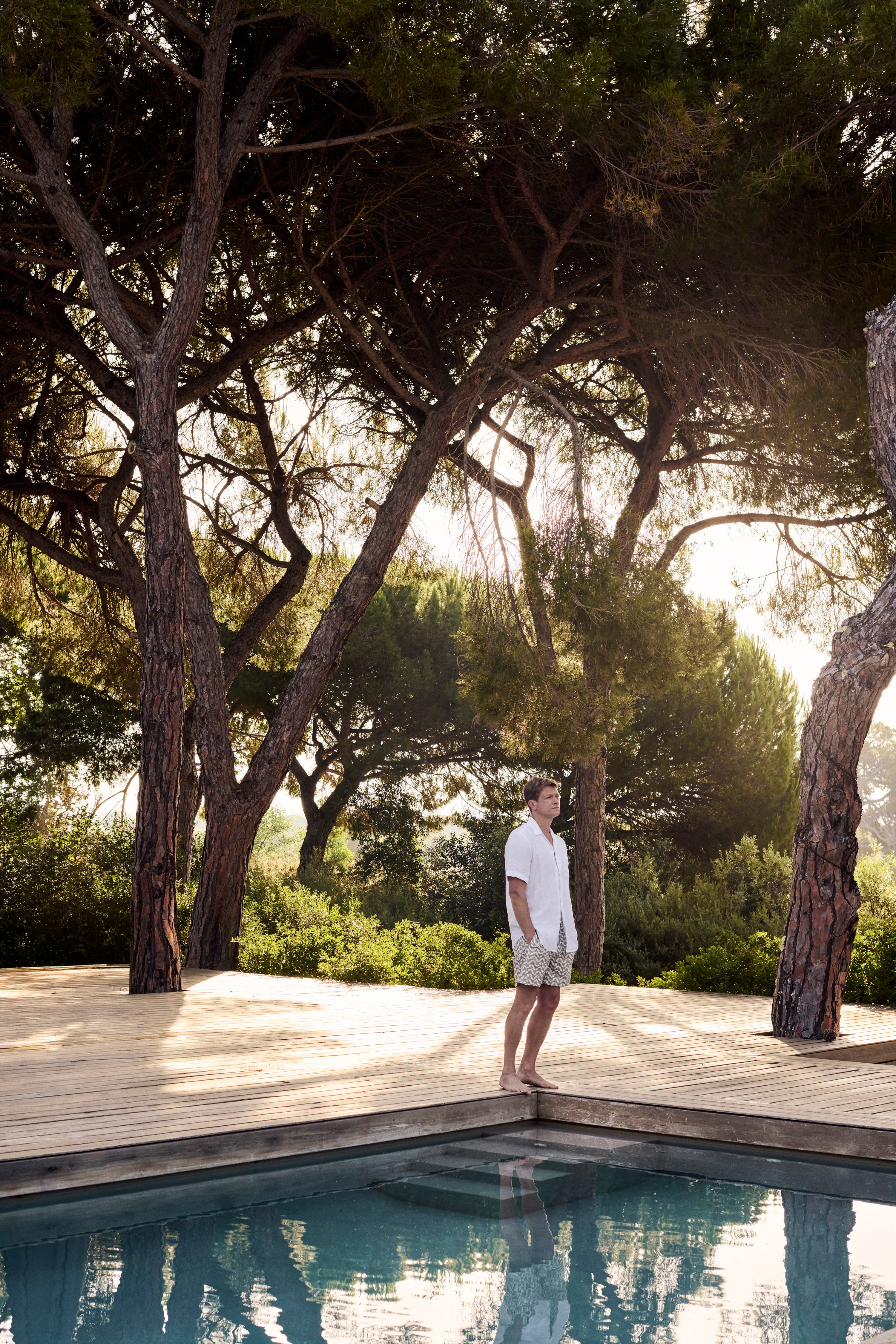 Ed Speleers in a white short-sleeve polo shirt and light beige shorts stands on the edge of a swimming pool beside a wooden deck, surrounded by tall trees in a sunny outdoor setting.