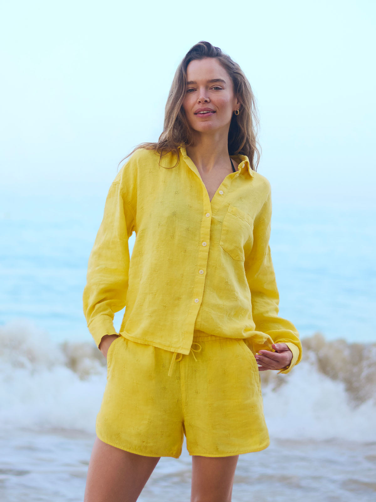 Woman in a yellow linen outfit standing on a beach with ocean waves in the background